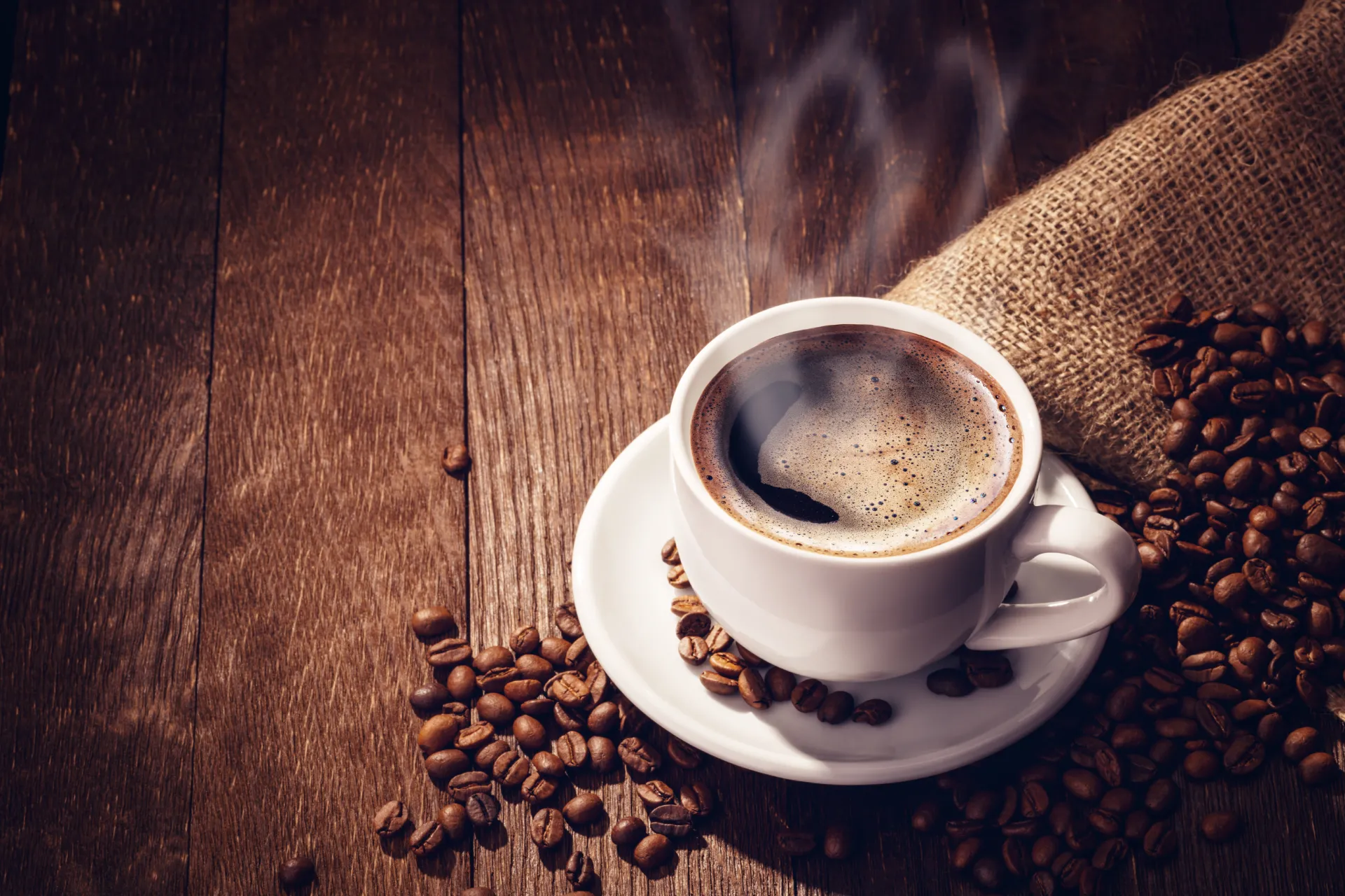 Konditorei Michael Baumhackl - Tasse mit frischem Kaffee auf Holztisch, Kaffebohnen drumherum